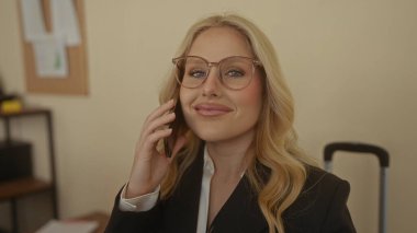 Modern bir ofiste telefonla konuşan, gülümseyen, gözlüklü, iş elbiseli, mutlu bir ifadeyle iç içe geçen, güven ve iletişimi vurgulayan sarışın bir kadın..