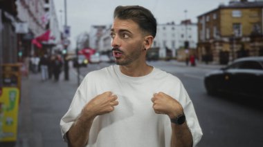 Beyaz tişörtlü, geniş gözlü adam iki elini de kendinle meşgul bir caddede gösteriyor..