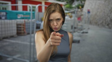 Gri kolsuz bluzlu genç bir kadın inşaat alanındaki iskelede parmağını kameraya doğrultuyor..