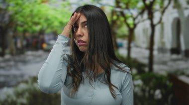 Bir sokakta genç bir İspanyol kadın, yorgun görünüyor ve alnına dokunuyor, yeşil ağaçlar ve arka planda bulanık binalar, açık mavi bir gömlek giyiyor.