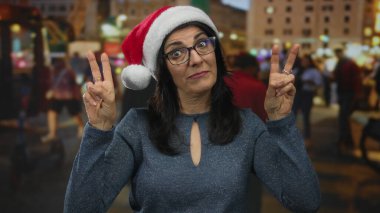 Orta yaşlı İspanyol kadın Noel Baba şapkası takıyor, geceleri işlek caddede parmak işaretleri yapıyor, şüphecilik.