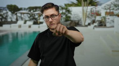Sakallı genç bir adam otelin havuzunda, sarp bir tatil köyü bahçesiyle çevrili, siyah bir gömlek ve gözlük takıyor, saygısız bir hareket sergiliyor, rahatlık ve rahatlama vurguluyor..