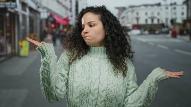Genç, kıvırcık saçlı, yeşil kazaklı, hareketli şehir caddesinde kafa karışıklığı yaşayan İspanyol bir kadın..