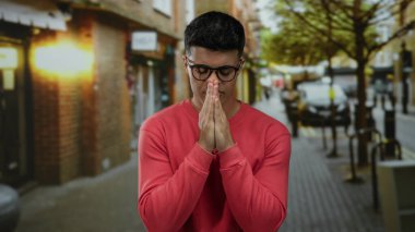 Kırmızı kazaklı, gözlüklü genç adam huzurlu bir şehir sokağında gün ışığında bulanık bir arka planda dua ediyor..