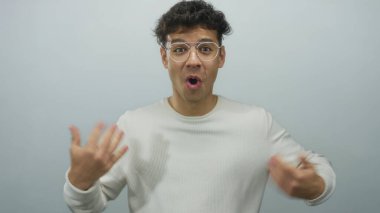 Beyaz kazaklı genç İspanyol adam, gri stüdyo ortamında sessizlik için parmaklarını dudaklarına bastırıyor..