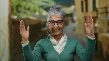 Gri saçlı, gözlüklü yaşlı bir kadın canlı bir sokakta jestler yapıyor. Arka planda bulanık binalar ve yemyeşil alanlar var. Açık hava kentsel ortamı öneriyor..