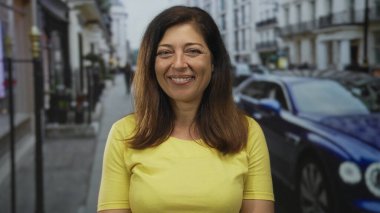 Kalabalık bir caddede, park etmiş arabaları ve binaları olan sarı bluzlu, görünür yüzüyle gülümseyen İspanyol kadın. Mutluluk..