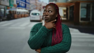 Şehir caddesinde kendine güvenen bir kadın yeşil süveter ve pembe örgüler takıyor. Etrafı şehir hayatı ve araçlarla çevrili. Açık havada güven ve şehir enerjisi taşıyor..