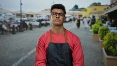 Gözlüklü ve önlüklü genç adam şehir caddesinde açık hava kafeleri ve canlı bir atmosferi olan güvenli bir şekilde duruyor..