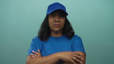 Mavi şapkalı ve mavi tişörtlü Afro-Amerikalı kadın kollarını kavuşturuyor ve stüdyoda ciddi bir ifade takınıyor..