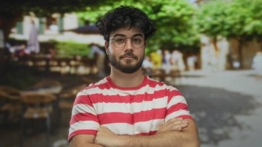 Gözlük takan sakallı genç adam güvenli bir şekilde ayakta duruyor. Arka planı bulanık, hareketli ve benzersiz bir sokak manzarası ve eşsiz bir stili olan hareketli bir sokakta kesişiyor..