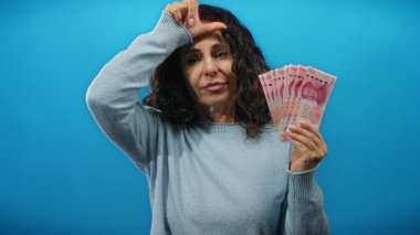 Orta yaşlı bir kadın, Çin Yuan banknotlarını mavi arka planda tutarak, duvarın içeriğinden izole edilmiş, kültürel ve finansal temalar yakalayan, kaybeden tabelalarını mizahi bir şekilde ifade ediyor..