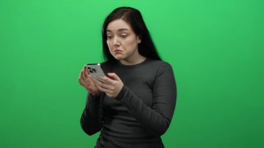 Akıllı telefonu parlak yeşil bir arka plana karşı kullanırken kaşlarını çatan kadın, hayal kırıklığını gösteriyor ve günlük kıyafetlere odaklanıyor, teknolojiyi ve ifadeyi ön plana çıkarıyor..
