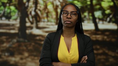 Kıvırcık saçlı bir kadın parkta kolları çapraz duruyor ve ciddi bir ifade takınıyor gözlük ve sarı bluz giyiyor..