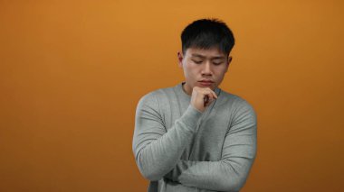 Genç adam, izole edilmiş turuncu bir duvara karşı düşünceli bir ifadeyle düşünüyor. Düşünce ve yansıma anı sergiliyor..