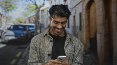 Genç İspanyol adam akıllı telefon tutuyor ve işlek bir şehir caddesinde gülümsüyor; anlık dijital bağlantı keyfi.