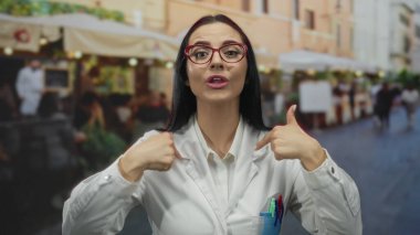 Laboratuvar önlüklü kadın, arka planda açık hava ortamında genç bilim adamını sergilemek için yoğun bir sokakta enerjik bir şekilde sesleniyor.