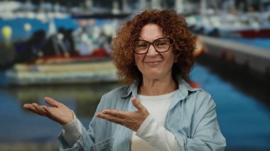 Orta yaşlı İspanyol kadın deniz kıyısında teknelerle el ele tutuşuyor, gözlük ve günlük gömlek giyiyor, açık hava ortamında bir merak ve nişan duygusu gösteriyor..