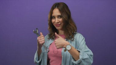 İngiliz anahtarı tutan İspanyol kadın, canlı mor bir arkaplan karşısında gülümseyerek, modern izole bir çevrede güven ve beceri ifade ediyor..