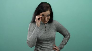 Gözlüklü bir kadın yeşil bir arka plan üzerinde gri bir kazak giyerek, modern gündelik tarzını ve düşünceli ifadesini özetliyor..