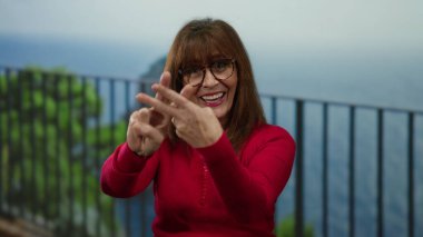 Kırmızı kazaklı İspanyol kadın hashtag jesti yapıyor. Okyanusun kenarında manzaralı bir arka plan gibi mutlu bir şekilde duruyor..