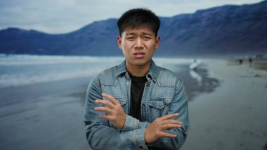 Sahilde genç bir adam kot ceketli parmaklarıyla okyanusa ve arka planda dağlara doğru hareket ediyor.