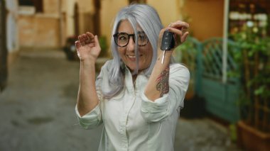 Son sınıf öğrencisi, gri saçlı, elinde araba anahtarlarıyla dışarıda güzel bir sokakta el sallayan, gözlüklü ve gülümseyen, neşeli bir an olduğunu öne süren bir kadın..