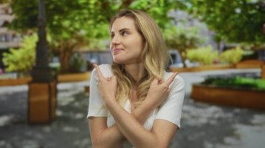 Parktaki bir kadın, yapraklı ağaçların altında dikilirken iki tarafı da çapraz kollarla işaret ederek doğal yeşil bir ortamda karar ya da yön öneriyor..