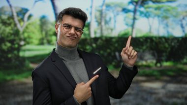 Açık hava parkında gülümseyen İspanyol bir adam gözlüklü ve günlük ceketli, kendine güvenen ve doğal çevrede çekici bir tavır sergiliyor.