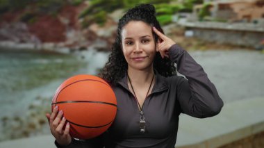 Plaj sahilinde basketbol oynayan bir kadın, boynunda ıslık çalan, açık gökyüzünün altında rahat ve düşünceli bir ifade sergiliyor..