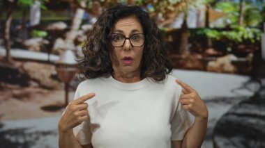 Orta yaşlı İspanyol kadın canlı yeşil parktaki göğüs göğse işaret ediyor; özgüven kimliği.