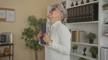 Kısa saçlı kadın ofis binasında tablet tutuyor ve yumruk sallıyor; başarı motivasyonu güven sevinci.