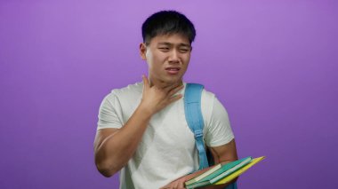 Beyaz gömlekli ve mavi çantalı genç bir adam mor arka planda kitap tutarken öksürüyor. Bu da soğuk algınlığı veya alerjisi olan bir öğrencinin izole edilmiş bir ortamda olduğunu gösteriyor..