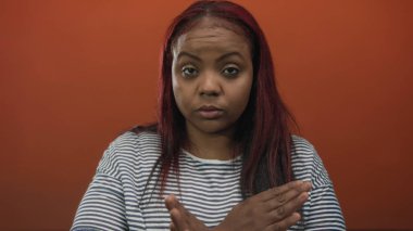 Canlı turuncu stüdyo duvarının önünde kollarını kavuşturan kadın gözleri kapalı çizgili bir gömlek giyiyor; meydan okuma sınırı.