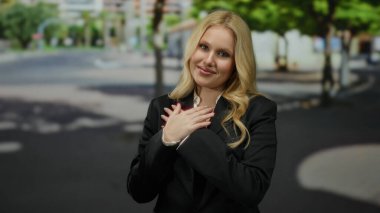 Şehirde minnettarlığını ifade eden bir kadın ağaçlar ve binalar, sarı saçlar, göğsünde eller, siyah bir takım elbise, güneşli bir gün, huzurlu bir atmosfer, gülümseme, dingin bir ruh hali.