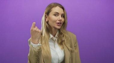 Ayrı bir mor arka plana karşı jest yapan, bej bir takım giyen, sohbet ya da etkileşim gösteren genç, kendine güvenen sarışın bir kişilik sergileyen bir kadın..