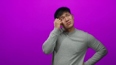 Genç Çinli bir adam, çenesinde elle pembe bir arka plana kafa yoruyor, düşünceli duygularını ifade ediyor, gündelik gri bir gömlek giyiyor, düşünceli bir ruh hali içinde..