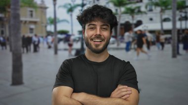 Palmiye ağaçları ve binaların yer aldığı kalabalık bir sokakta kollarını kavuşturup güvenle gülümseyen genç İspanyol adam..
