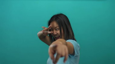 Çinli kadın gözünün yanındaki parmaklarıyla neşeli bir jest yapıyor, canlı yeşil bir duvara gülümsüyor, gençlik enerjisi ve güveni sergiliyor..