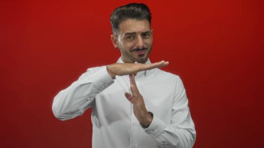 Beyaz gömlekli İspanyol genç adam kırmızı stüdyo ortamında el hareketleriyle ara veriyor..