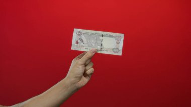 Kafkasyalı bir adam Birleşik Arap Emirlikleri 'ne bağlı Dirham Dirham banknotunu canlı kırmızı bir arka plana karşı tutuyor, izole ve odaklanmış bir şekilde para gösteriyor..