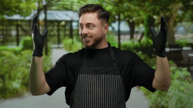 Önlük ve eldiven giyen genç adam park ortamında jest yapıyor, doğa ve hizmet karışımını yakalıyor, davetkar yeşil bir ortamda profesyonel konukseverlik gösteriyor..