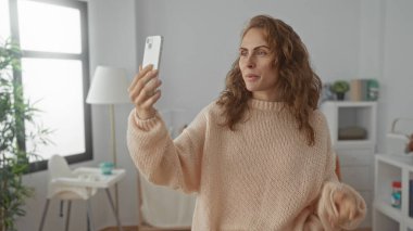 Bebek beşiği ve modern dekoru olan rahat bir yatak odasında, kazak ve kot pantolon giyen, akıllı telefon tutarken duygularını ifade eden bir kadın videosu..