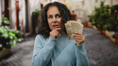 Güney Afrika Rand 'i elinde tutan orta yaşlı bir kadın bir şehir caddesinde düşünceli bir şekilde ayakta duruyor..