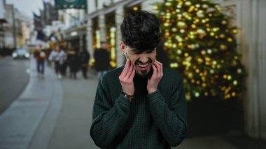 Şehir sokağında genç bir adam tatil sezonunda yüzü ağrıyan bir diş ağrısı çekiyor ve arka planda bir Noel ağacı var..