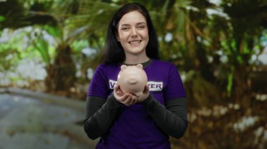 Yeşil parkta elinde domuz kumbarasıyla gönüllü olarak çalışan, mor gömlek giyen, yardım, mutluluk ve toplum desteği taşıyan bir kadın..