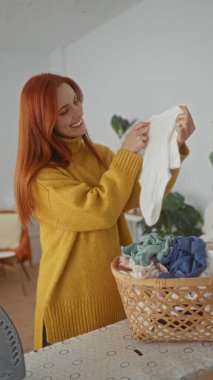 Evde rahat bir çamaşırhanede elbiselerini katlayan, kızıl saçlarını sergileyen ve sarı bir kazak giyen, yeni temizlenmiş çamaşır sepetleriyle çevrili bir kadın..