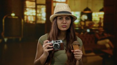 Dondurma ve kamera taşıyan bir kadın loş bir otel salonunda, hasır şapka takıyor, seyahat titreşimlerini yansıtıyor merak ve macera ile.