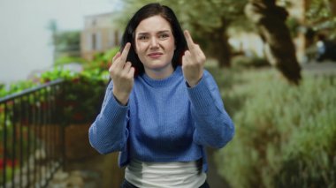 Yeşil bir parkta dikilirken orta parmaklarıyla jest yapan bir kadın, aydınlık bir günde açık havada yakalanan bir ortamda hayal kırıklığı ve tavır sergiliyor..