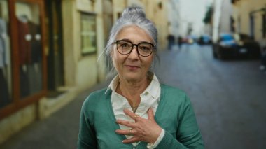 Gri saçlı ve gözlüklü yaşlı bir kadın şehir sokaklarında güvenle gülümsüyor, yeşil bir hırka giyiyor, şehirde sıcaklığını ve bilgeliğini iletiyor..